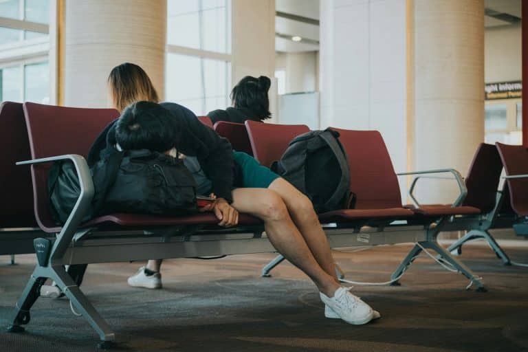 A man lies tired on the seats of an airport, his head resting on his bag. We can't see his face, only the top of his head. He is wearing green shorts, a black jacket and white trainers. Two other people are sitting in the chairs behind him.