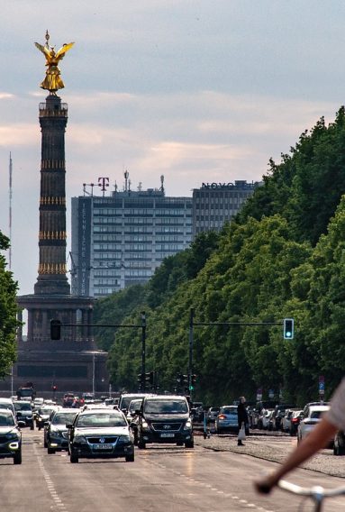 Photo of the Siegessäule in the centre of the Großer Tiergarten in Berlin, there is traffic around the square and many trees line the avenue.