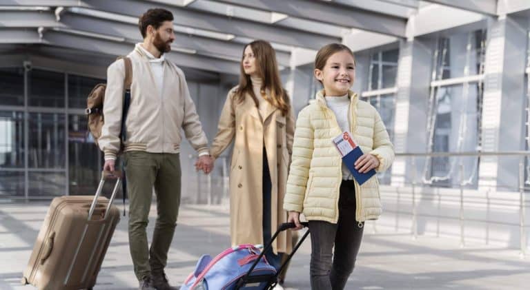 A family of three, a couple and their young daughter, are walking along an airport departure corridor. They are going on a trip. The couple walks at the back, holding hands. They are wearing beige clothes and the man is pulling a suitcase of the same colour. The smiling little girl is walking at the front, pulling her blue suitcase. She is holding her passport and boarding pass in her hand.