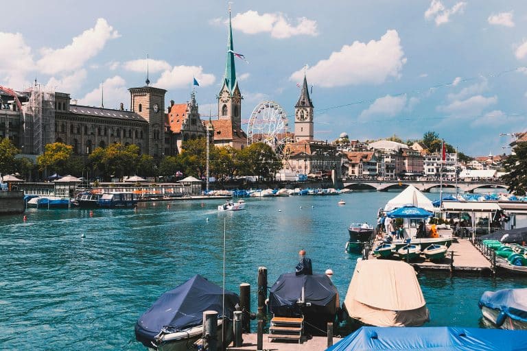 Photo of the beautiful city of Zurich in Switzerland. Pleasure boats and tourist boats are moored along the quays. Many tourists are visiting the city and taking photos on the bridge. There is a large Ferris wheel installed on one of the riverbanks.