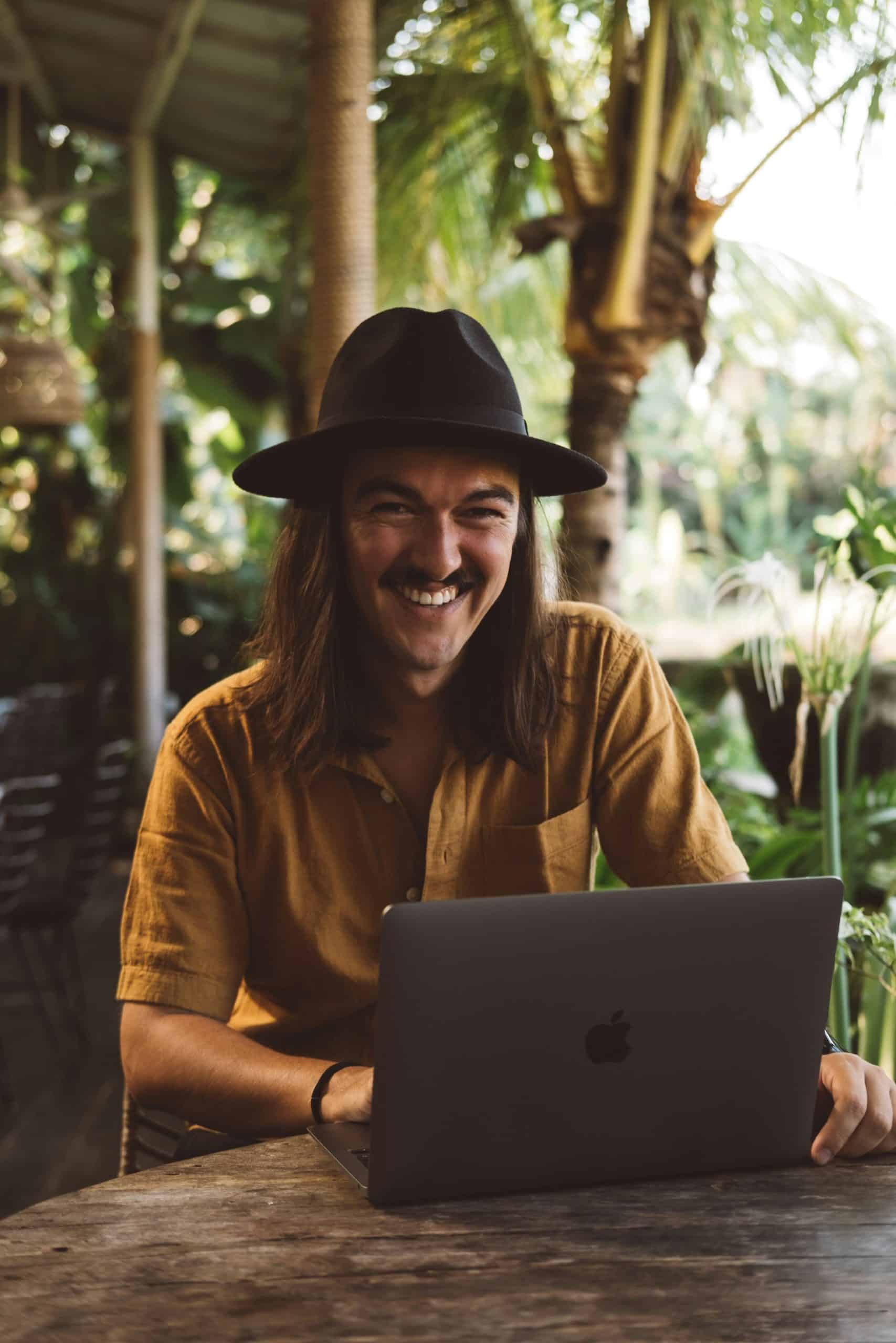 A man with a moustache and wearing a black hat smiles at the camera. He is wearing a mustard yellow shirt. He is sitting at a table on the terrace of a café and working on his laptop. He is in a tropical country, judging by the landscape and palm trees behind him.