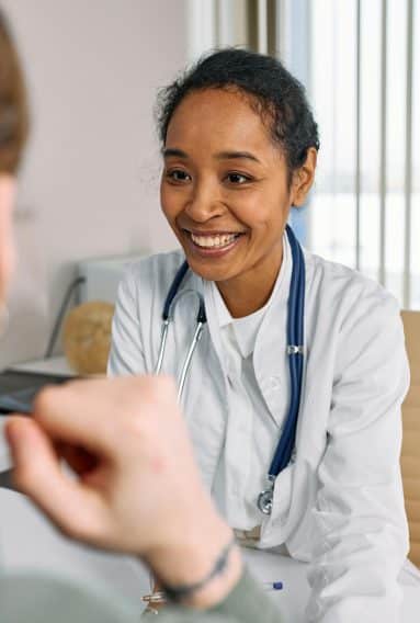 A doctor smiles at her patient with her stethoscope around her neck. The doctor is wearing a white coat and her hair is tied back.
