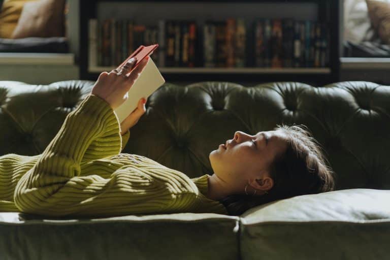 A young woman in an olive green jumper is lying on a sofa of the same colour, close to khaki. She is holding a red book in her hands and appears to be deep in thought.