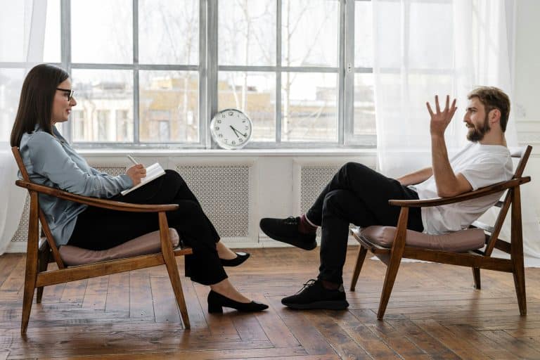 A woman and a man, both wearing glasses, are sitting facing each other on chairs in front of large windows in a room, talking together in therapy. The woman is taking notes in a notebook, suggesting that she is the coach, while the man is speaking; he is undoubtedly her patient.