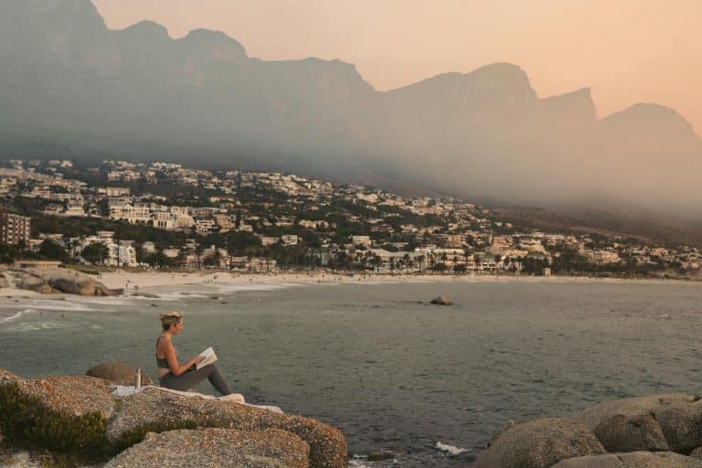 A young blonde woman in yoga attire is sitting on a towel on a cliff, reading a book resting on her lap. She is facing the landscape of Cape Town beach in South Africa.