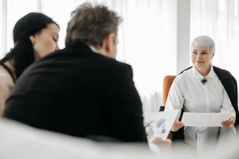 A woman with short white hair, dressed elegantly and distinguished, welcomes a man and a woman into her office. The setting is simple and white, and they are seated on brown leather chairs.