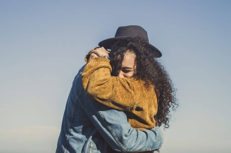 A woman in a yellow jumper with long brown curly hair embraces a man wearing a hat and a corduroy jacket. The woman looks happy. The background of the photo is a large blue sky.