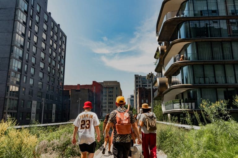 Three young men with an urban, trendy style are walking outside in a park surrounded by tall buildings. The weather is nice and clear.
