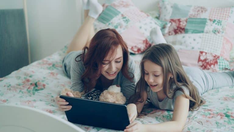 A mother and her daughter are lying on her bed and looking at a tablet together. The mother is holding a teddy bear in her arms. They are both dressed in grey and wearing white socks. They are smiling and in a good mood.