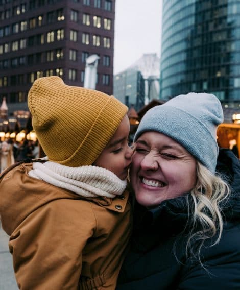 A small child is kissing his mum's cheek in winter time, in a christmas market located in the centre of a urban city in Germany.