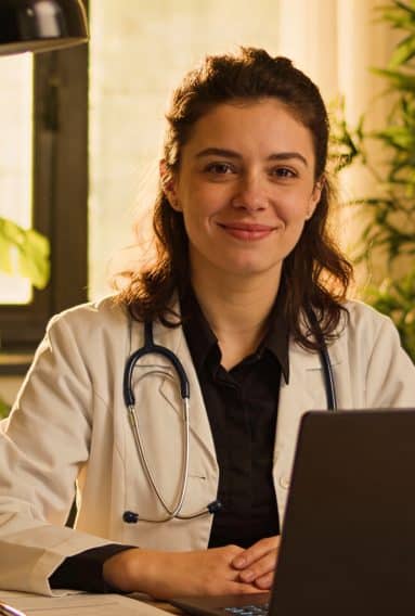 Healthcare professional wearing a white coat and stethoscope, seated at a desk with a laptop in a warmly lit room filled with plants.