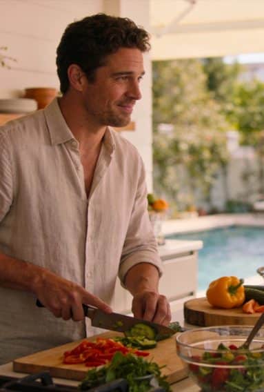 Person slicing vegetables on a wooden cutting board in an outdoor kitchen next to a swimming pool, surrounded by fresh produce