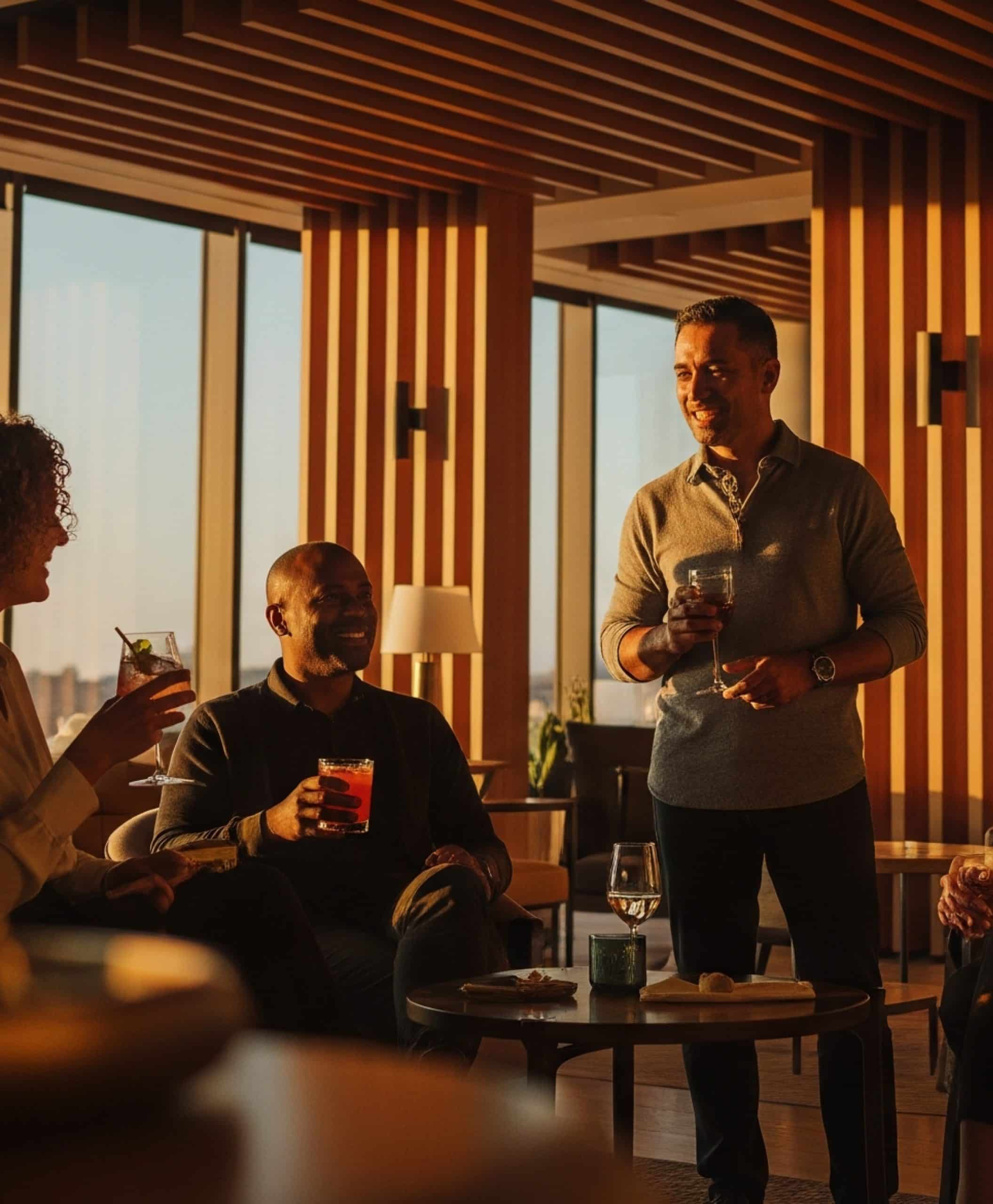 Several people are gathered in a modern lounge area with tall wooden panels and large windows letting in warm sunset light. One person is standing while holding a drink, and the others are seated with drinks in hand around small circular tables. The scene has a relaxed, upscale atmosphere.