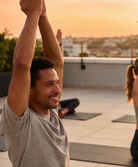 Person practicing yoga on a rooftop at sunset, sitting on a mat with arms raised overhead. Other participants and city buildings are visible in the background.