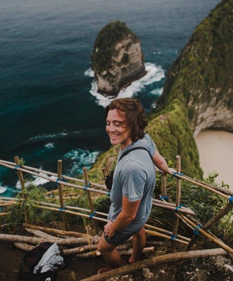 A person stands on a narrow, rustic wooden pathway built along a cliffside overlooking the ocean. Below, waves crash against tall, steep rock formations covered in greenery. A sandy beach sits in a cove to the right. The person is wearing casual outdoor clothing and holds onto the bamboo railing while looking toward the dramatic coastal landscape.