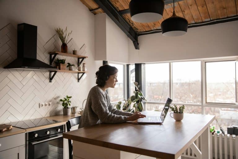 A person sits at a wooden kitchen island using a laptop in a bright, modern kitchen with large windows, indoor plants, and exposed wooden ceiling beams.