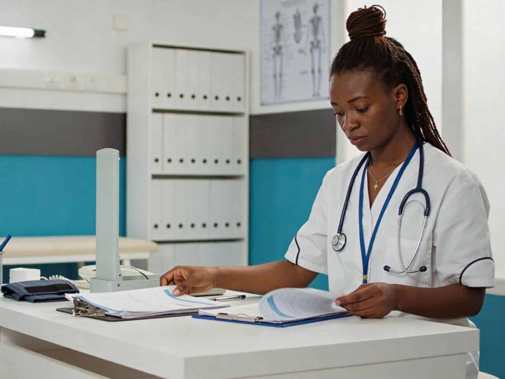 Medical practitioner sitting at a desk in a clinic, reviewing patient files with medical equipment and charts visible in the background