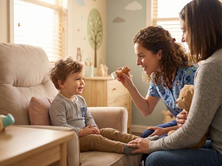 A child sitting on a small sofa in a pediatric examination room while a healthcare professional gently checks their eyes with a small light. An adult sits nearby holding a teddy bear, offering comfort.
